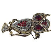 Broche colgante  buho de metal dorado y brillantes de strass en color rojo. Trasera en color oro viejo con alfiler y seguro. Además del alfiler habitual, lleva una pieza que permite usarlo como colgante. Mide 6 cm de largo x 3 cm de ancho.